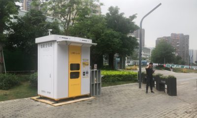 A yellow-and-white Meituan pickup kiosk in front of trees. A man is standing nearby.